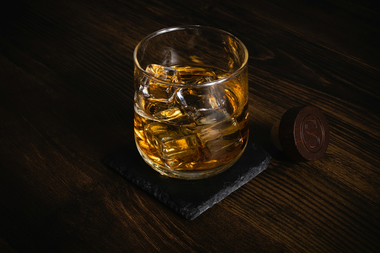 A glass of whisky with ice beside a monogrammed wooden cork stopper on a dark table, set for a considered gift occasion.