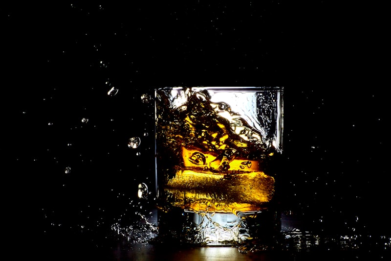 A glass of whisky mid-splash as ice cubes drop in, with droplets suspended against a black background.