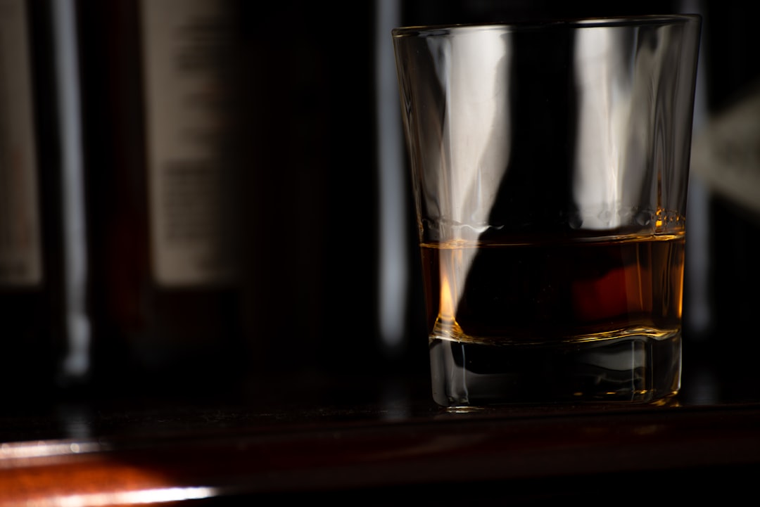 A glass of whisky catching light on a dark bar counter, with bottles blurred in the background