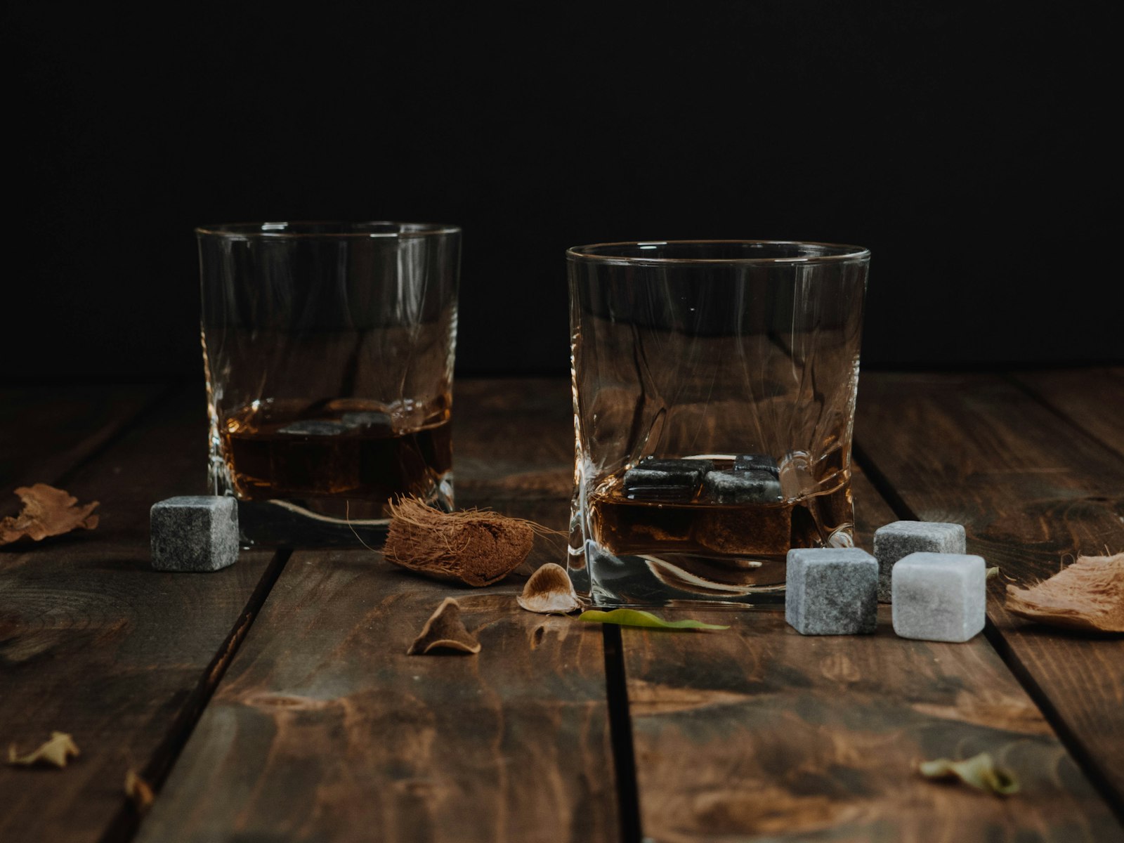 Two whisky glasses with ice cubes and whisky stones on a dark wooden table.