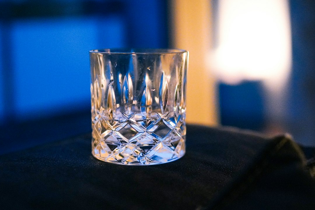 Empty cut-crystal whisky tumbler on a dark fabric surface with blurred blue and warm lights behind