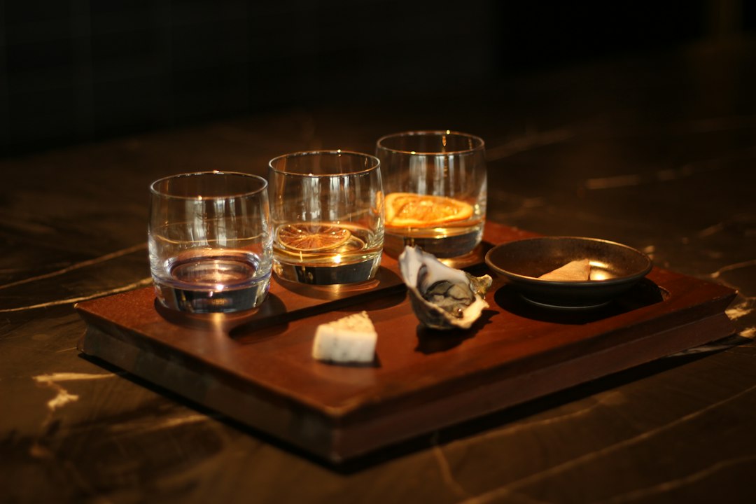 Three tumblers of whisky in a tasting flight on a wooden tray, with small food pairings, on a dark marble table